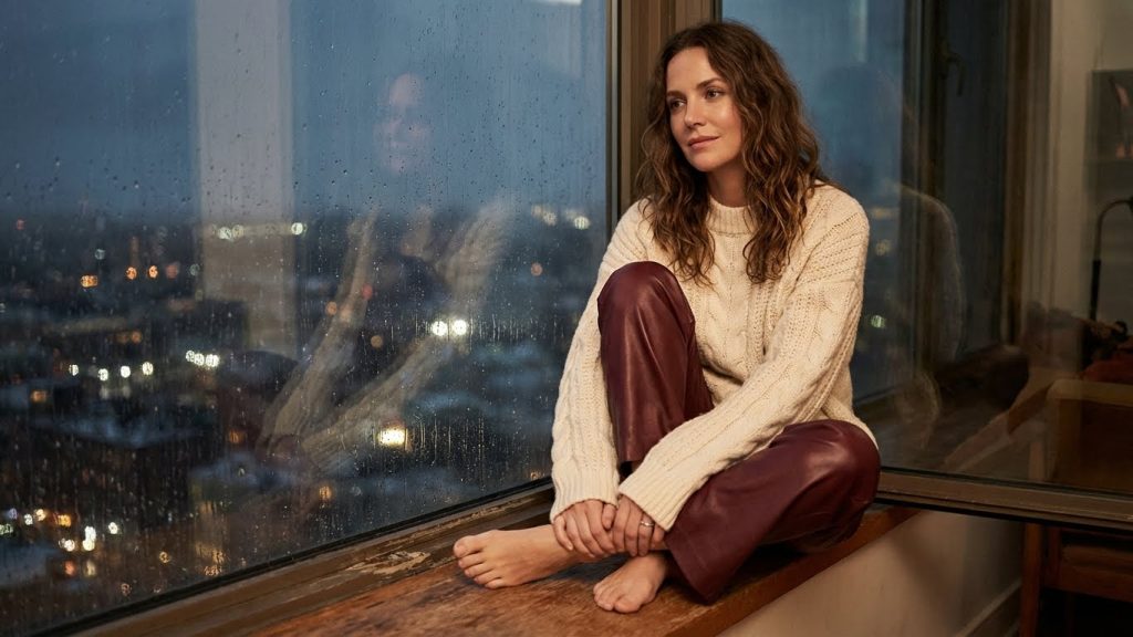 Woman in burgundy leather pants and cream sweater gazing out rainy window at city lights.