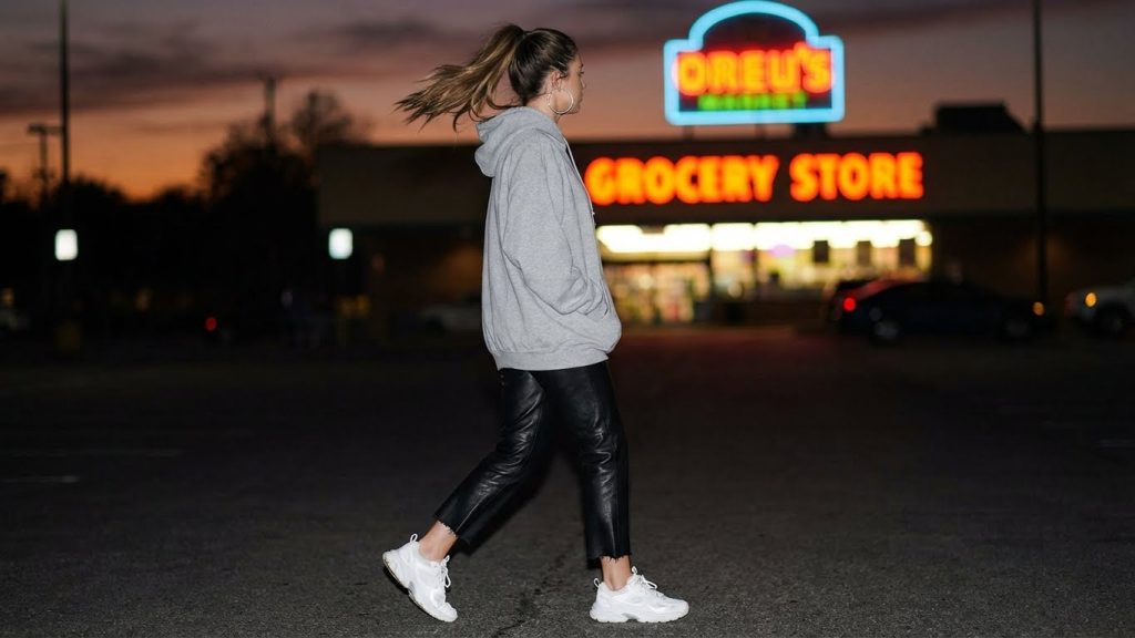 Woman in gray hoodie, cropped leather pants, and white sneakers walking at night.