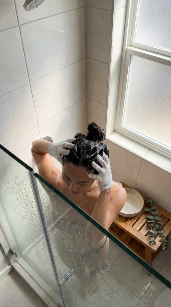 Woman in shower washing hair with scalp massage gloves.