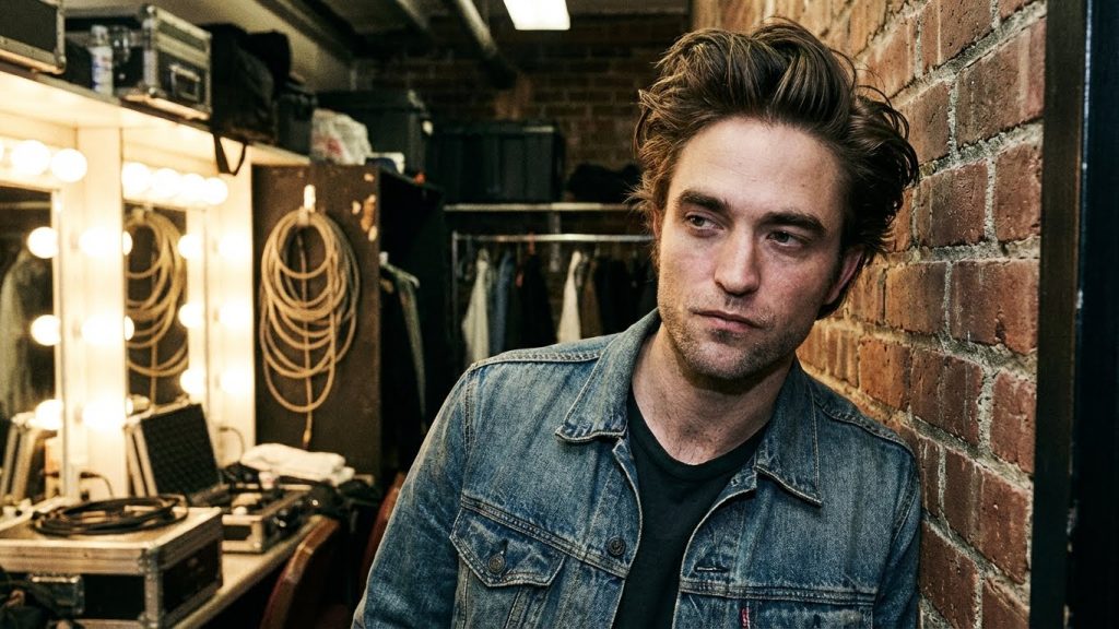 Robert Pattinson in a modern pompadour medium length hairstyle for men, leaning against a brick wall.