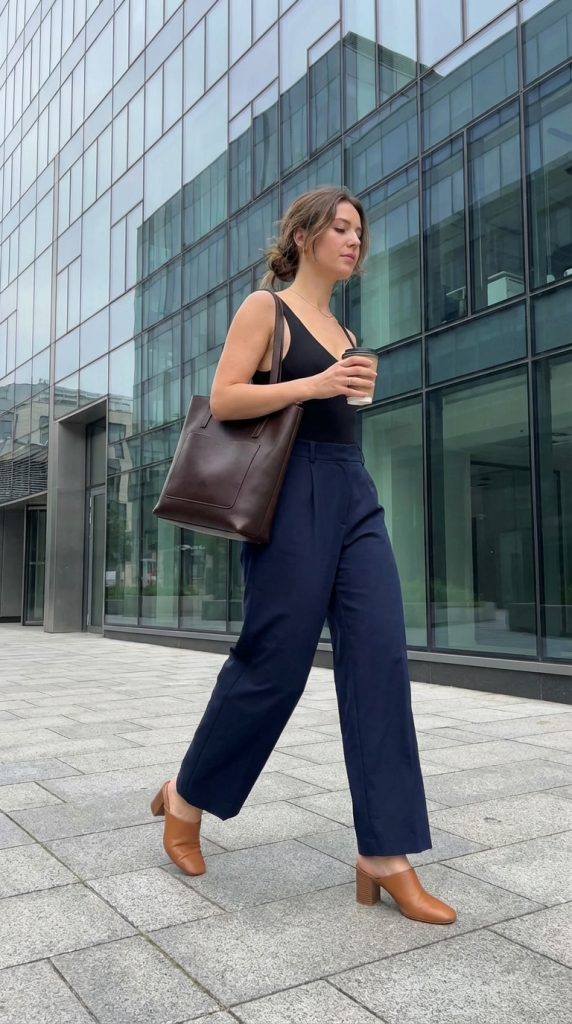 Woman in wide-leg trousers, black bodysuit, brown mules, and a tote bag walking past an office building.