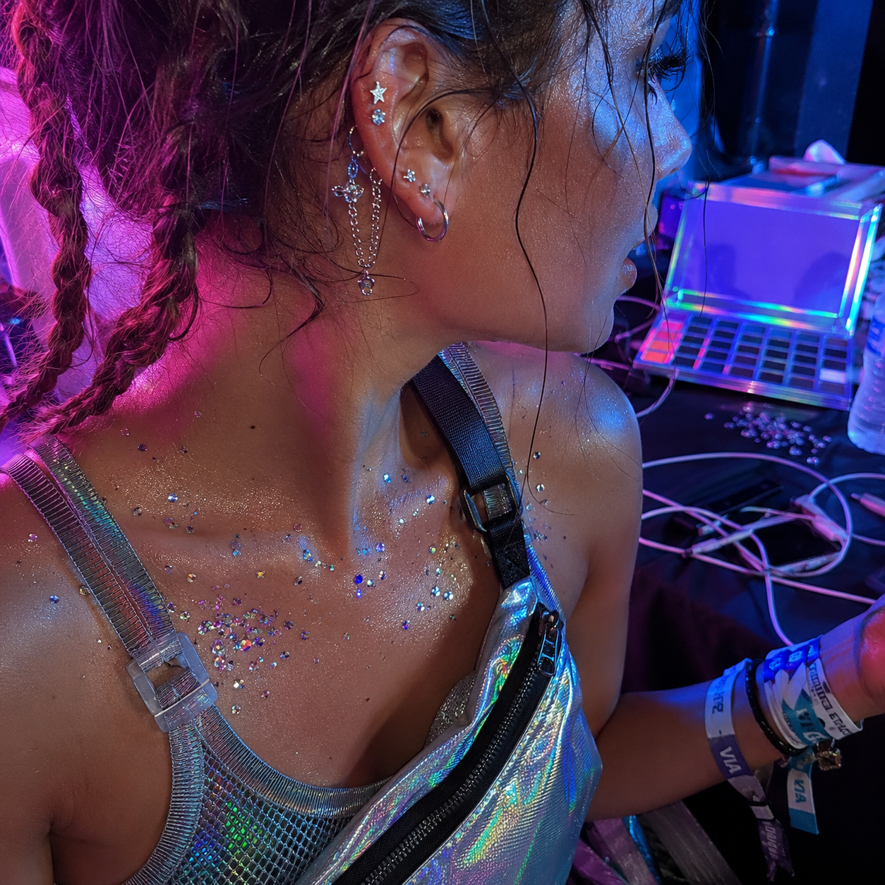 Side profile of person in neon light with glittery chest, multiple ear piercings, and holographic top beside laptop.