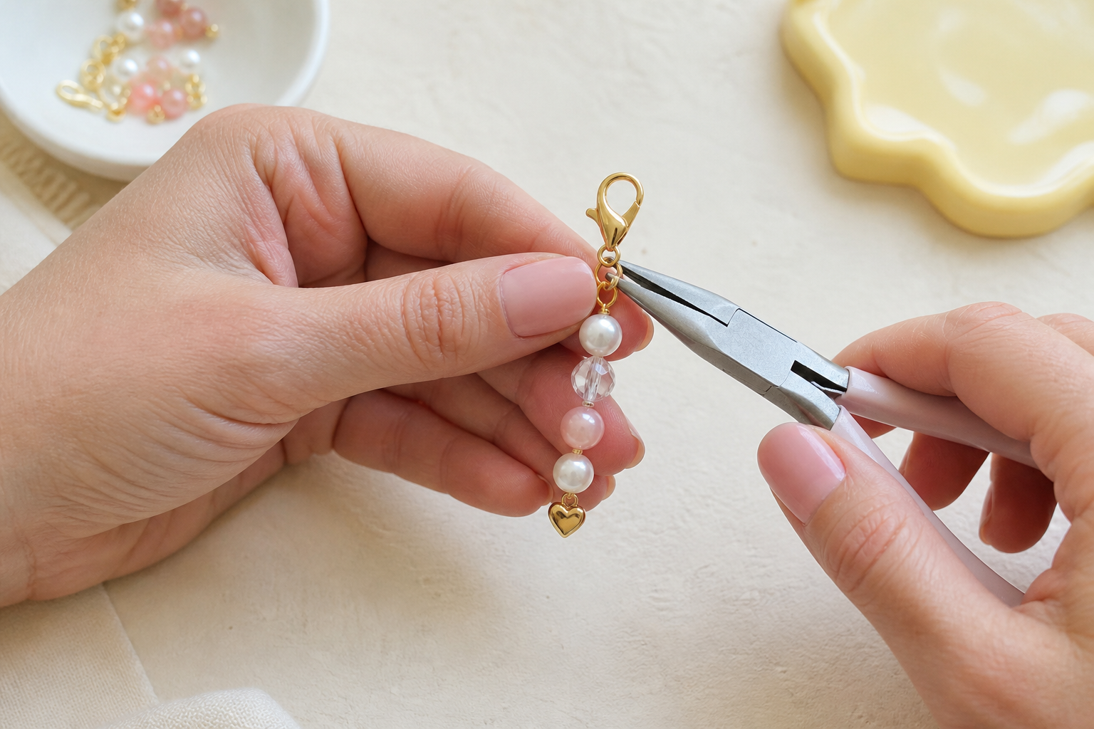 Hands use pliers to attach a gold lobster clasp to a beaded charm featuring pearls, a clear crystal, and a small gold heart.