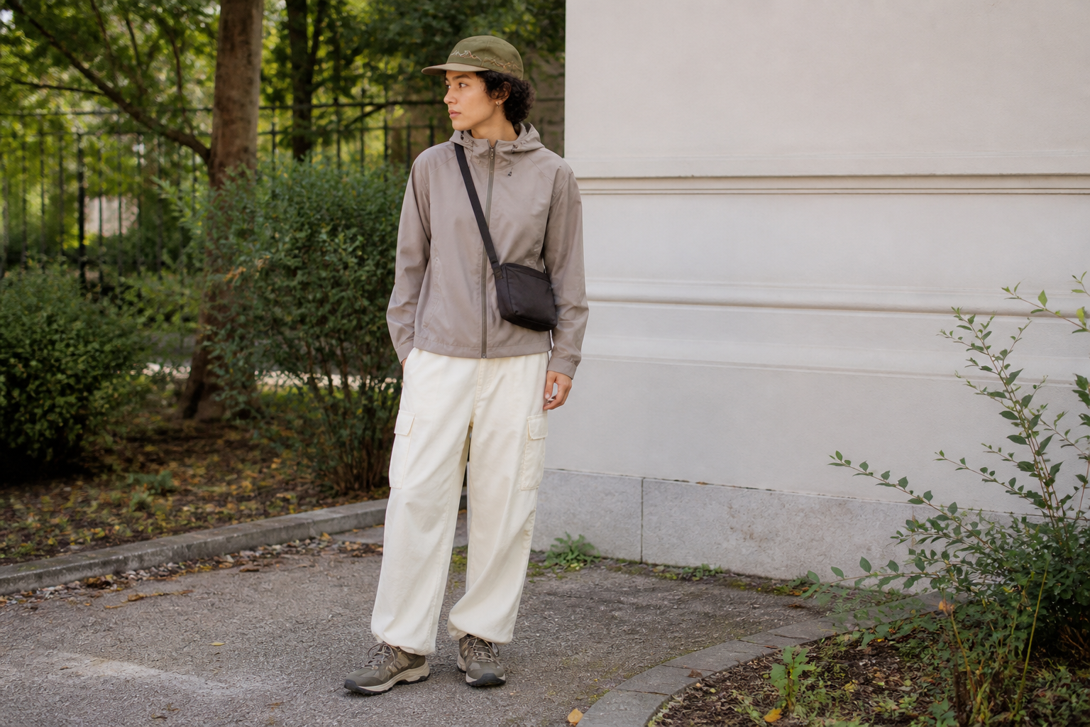 A person stands outdoors wearing an olive cap, taupe windbreaker, cream cargo pants, and grey sneakers, carrying a black crossbody bag against a white wall and greenery.