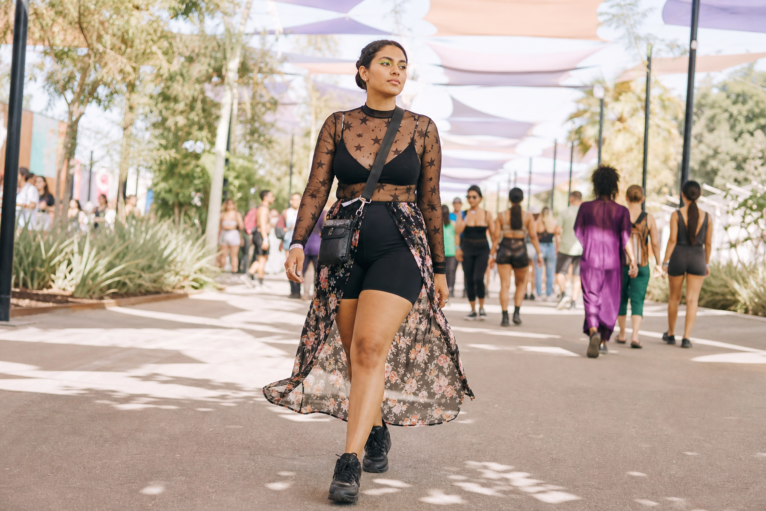 A woman with green eyeshadow walks forward wearing a black star-mesh top, biker shorts, floral duster coat, and sneakers. She carries a crossbody bag under a colorful canopy at an outdoor event.