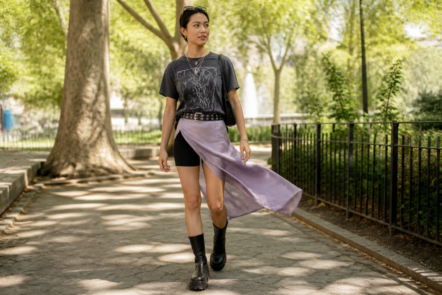 A woman walks on a park path wearing a graphic tee, black biker shorts, and a flowing lavender satin skirt over them, paired with chunky black boots and a studded belt.