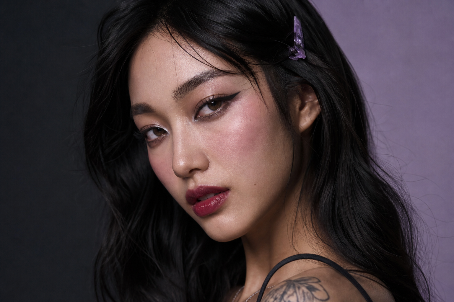 Close-up portrait of a woman with dark hair, winged eyeliner, and berry lips. She wears a purple butterfly hair clip and has a shoulder tattoo against a split black and purple background.