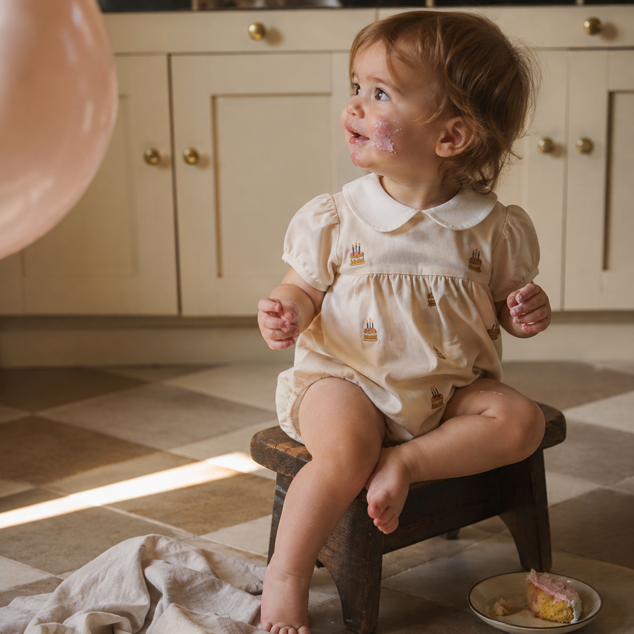 Toddler with pink frosting on face sits on a wooden stool wearing a cream romper with embroidered birthday cakes. A pink balloon floats nearby, and a slice of cake rests on the floor.