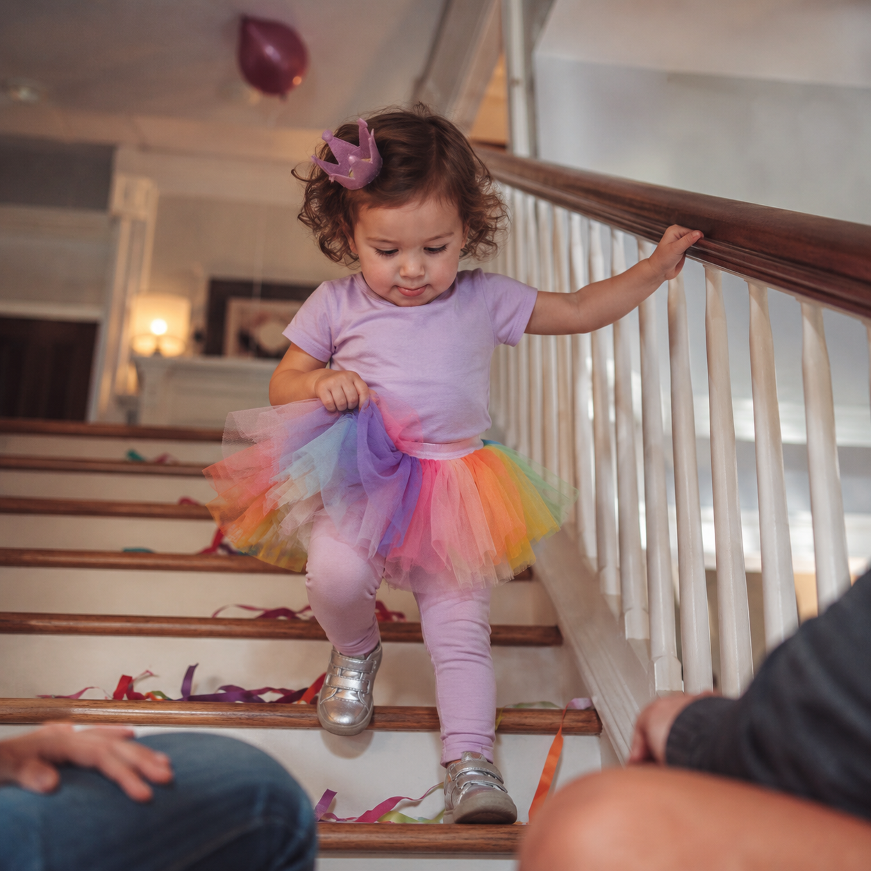 A toddler in a purple crown, rainbow tutu, and silver shoes carefully descends stairs scattered with party streamers, holding the wooden railing.