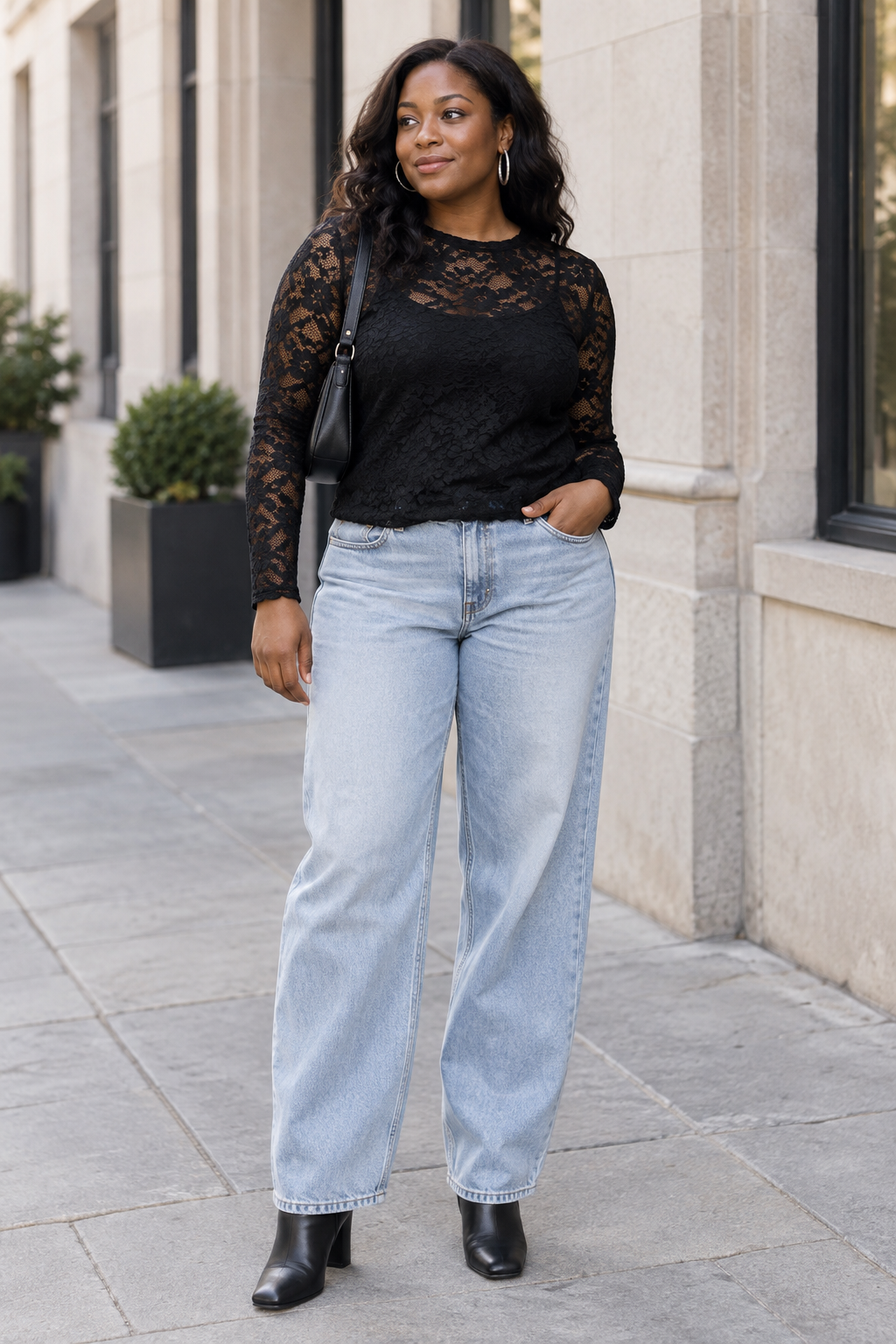 A woman stands outdoors wearing a black lace long-sleeve top, light wash wide-leg jeans, and black block-heel boots. She carries a small black shoulder bag and wears silver hoop earrings.
