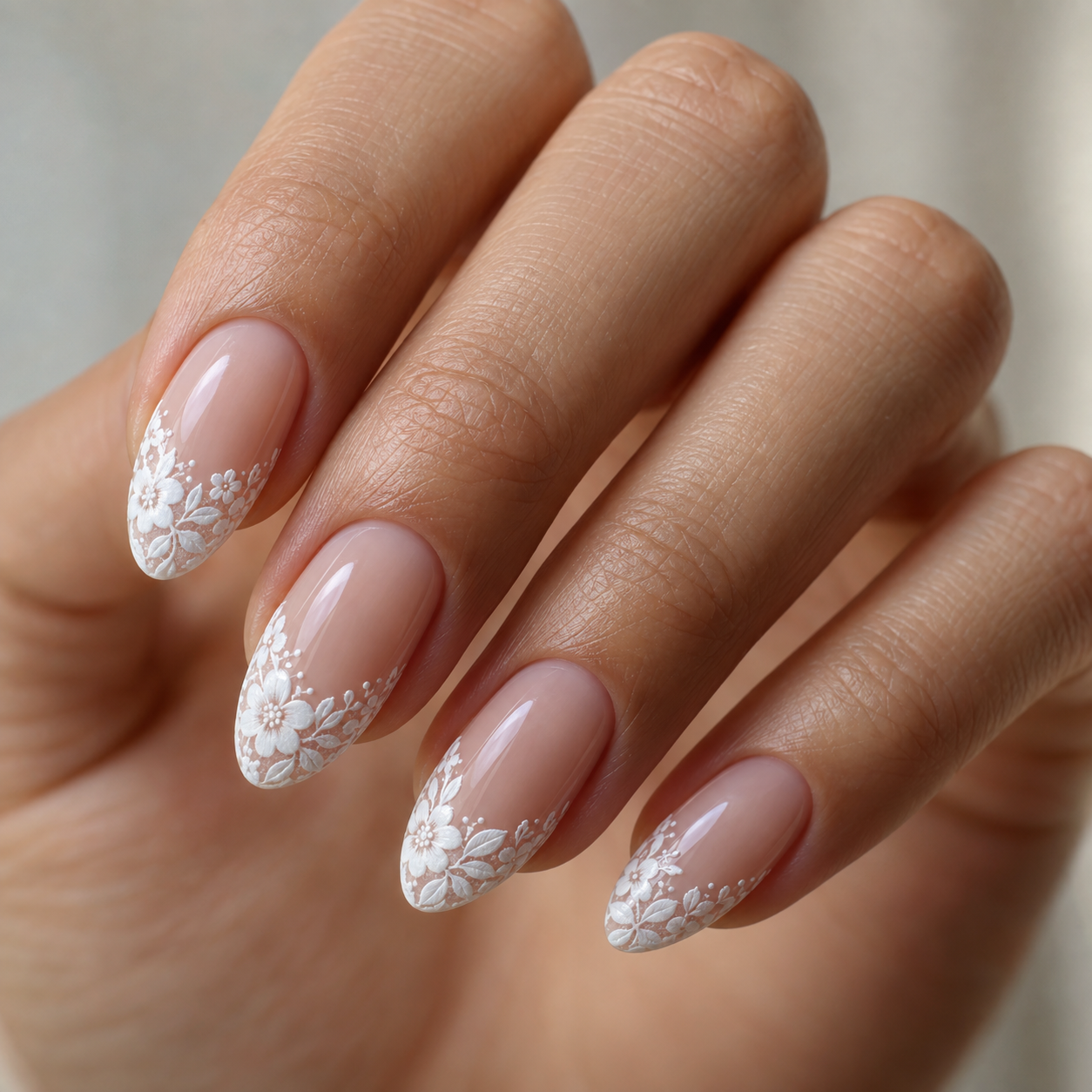 Almond nails with glossy nude base and intricate white floral lace detailing along the tips, featuring delicate flowers and leaves.