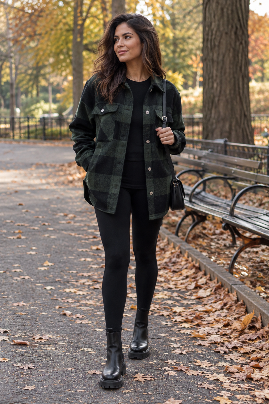Woman in a green and black plaid flannel shirt, black top, leggings, and chunky boots stands on a leaf-strewn park path near benches.
