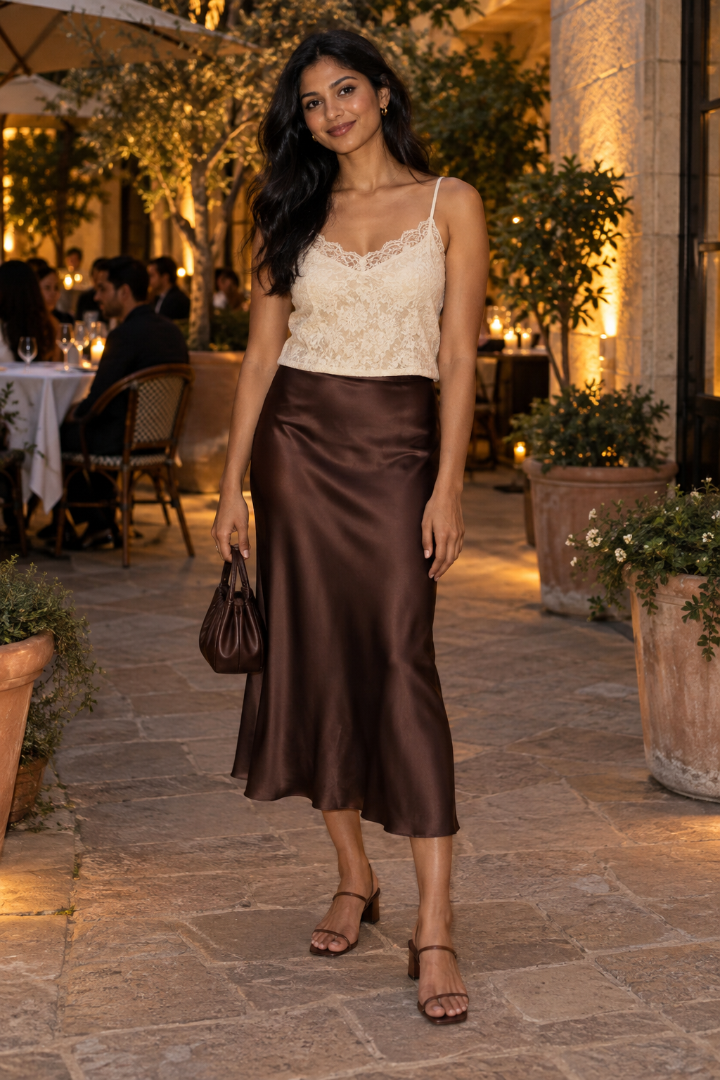 Woman in a cream lace camisole and brown satin midi skirt holding a small brown handbag, standing on a stone patio at an outdoor restaurant.