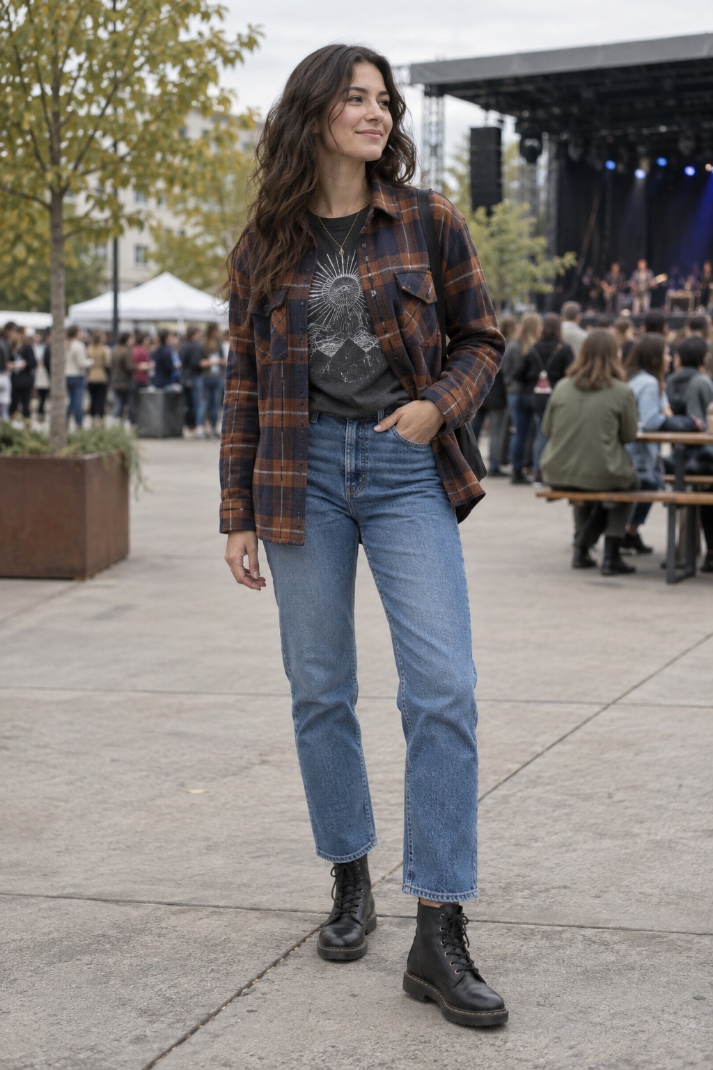 A woman with long dark hair wears an open brown plaid flannel over a graphic tee, blue jeans, and black combat boots at an outdoor concert venue.