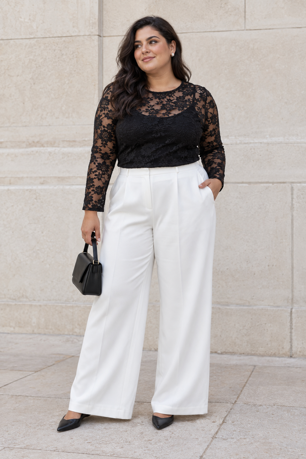 A woman stands against a stone wall wearing a black lace long-sleeve top, white wide-leg trousers, and black pointed-toe heels. She holds a small black handbag.