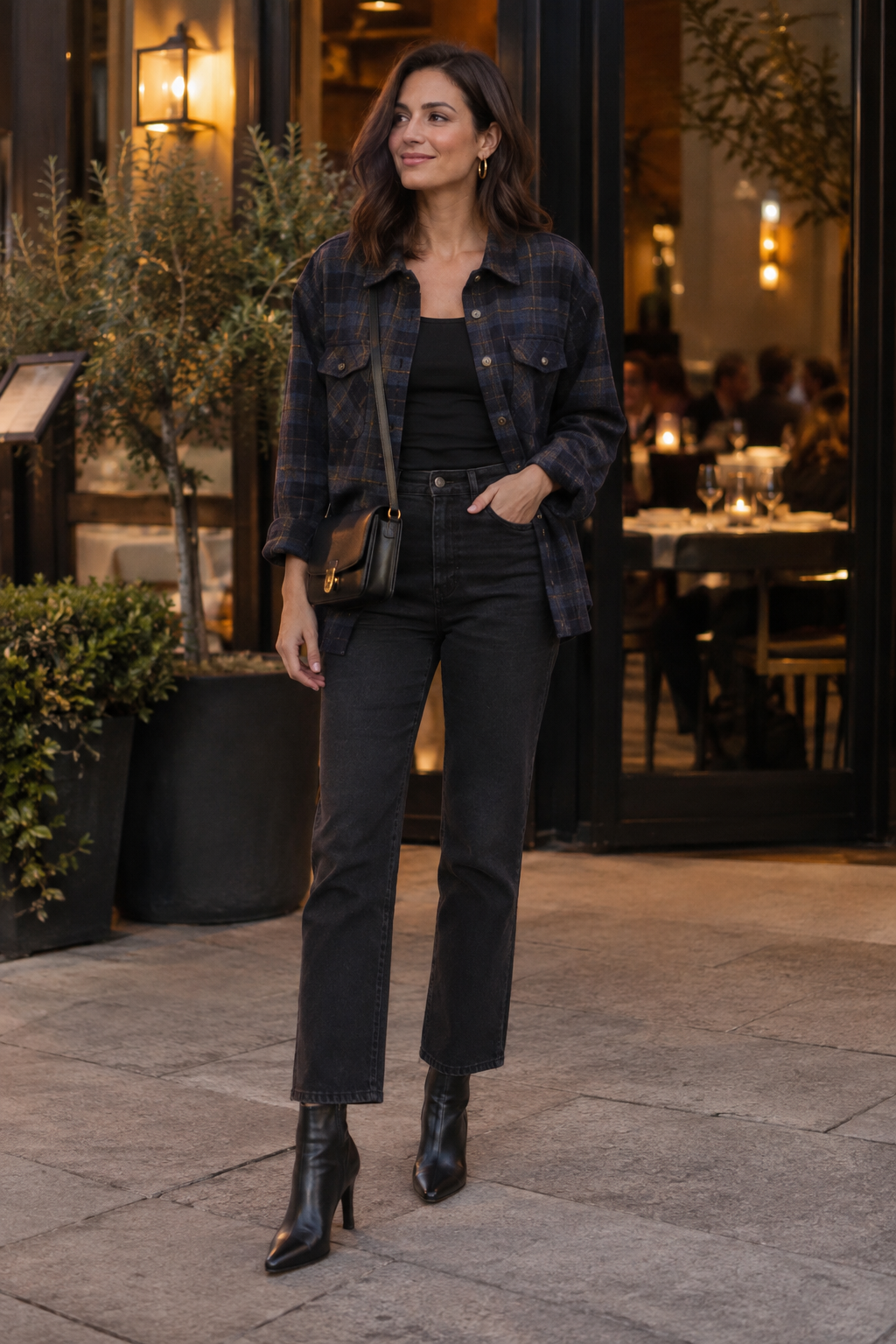 Woman in a blue plaid shirt, black top, dark straight-leg jeans, and black heeled boots stands outside a restaurant with warm lighting and potted plants.