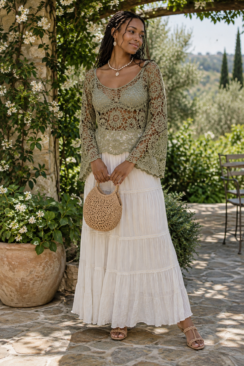 Woman in sage green crochet top and white tiered maxi skirt holding a woven straw bag on a stone patio with climbing white flowers.
