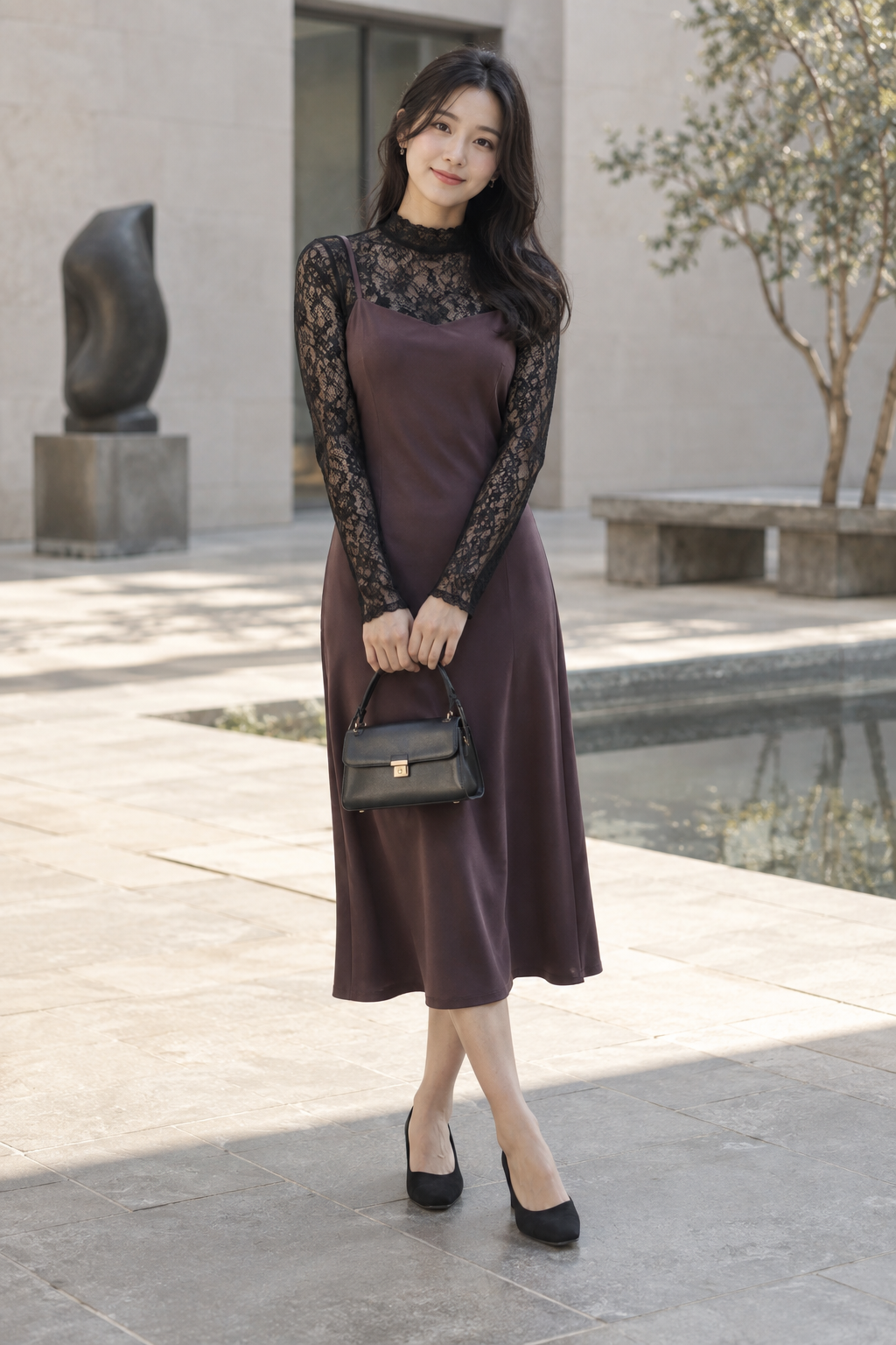 A woman stands outdoors wearing a plum satin slip dress layered over a black lace turtleneck. She holds a small black handbag and wears black heels, with a sculpture and water feature in the background.