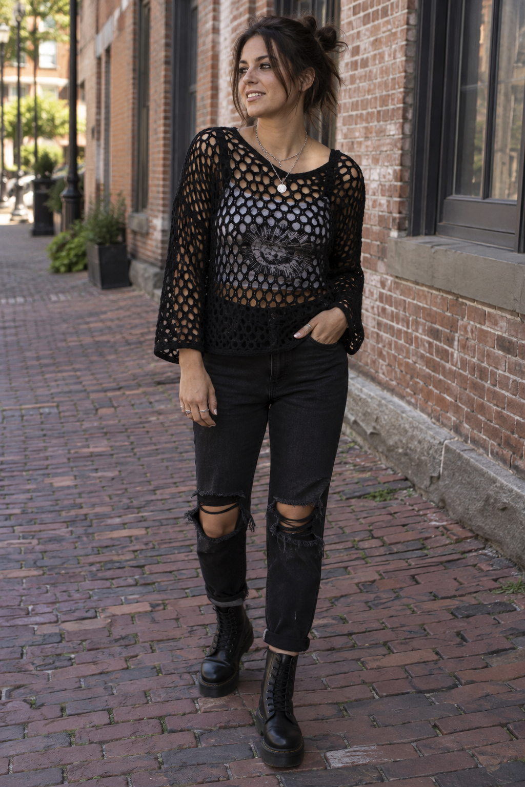 A woman stands on a brick sidewalk wearing a black fishnet sweater over a graphic tee, ripped black jeans, and black combat boots.