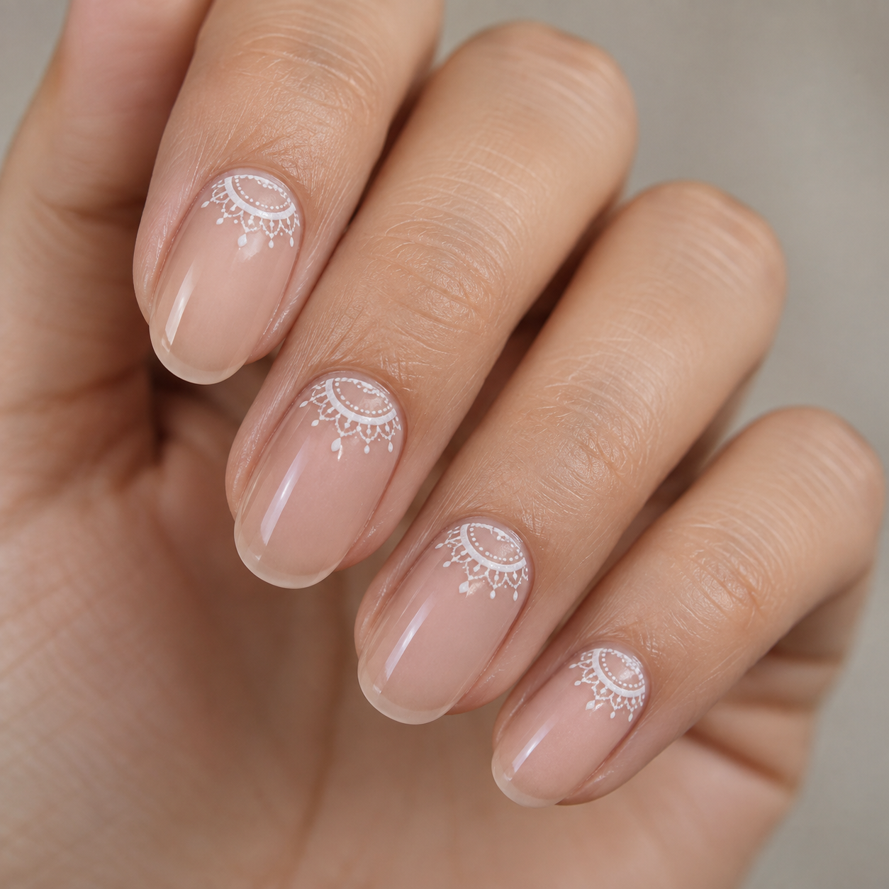 Nude manicure featuring delicate white lace or mandala-style art near the cuticles on four fingers.
