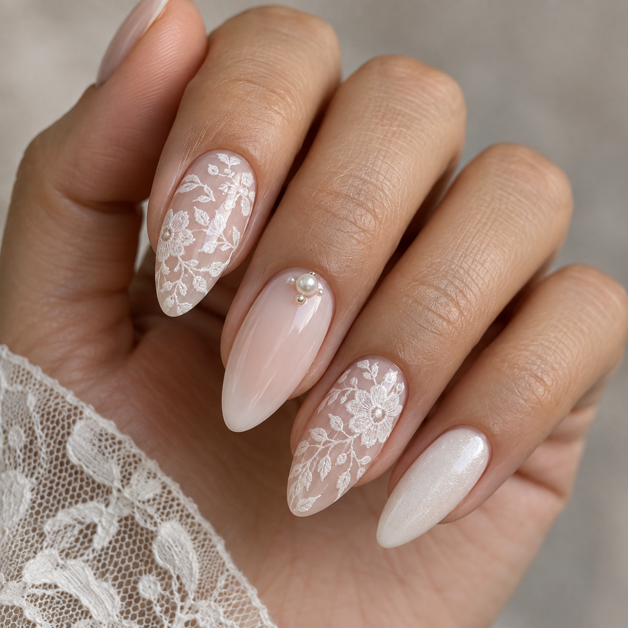 Almond nails with white lace floral patterns, a nude accent with pearls, and shimmery white polish. Hand rests near white lace fabric.