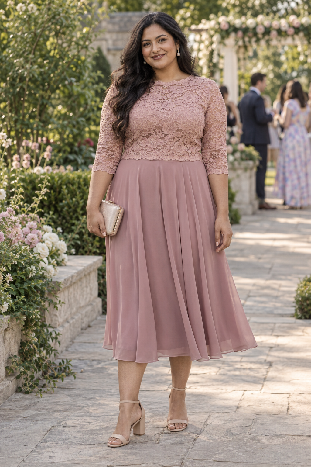 A woman stands on a stone path in a garden wearing a dusty rose lace top and matching pleated midi skirt. She holds a gold clutch and wears nude block heels.