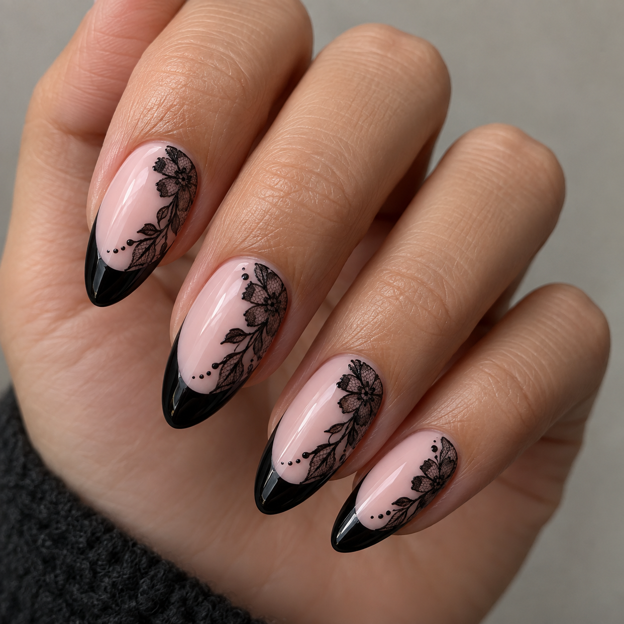 Almond nails feature a black French tip on a pale pink base, decorated with intricate black floral lace art and small dots along the smile line.