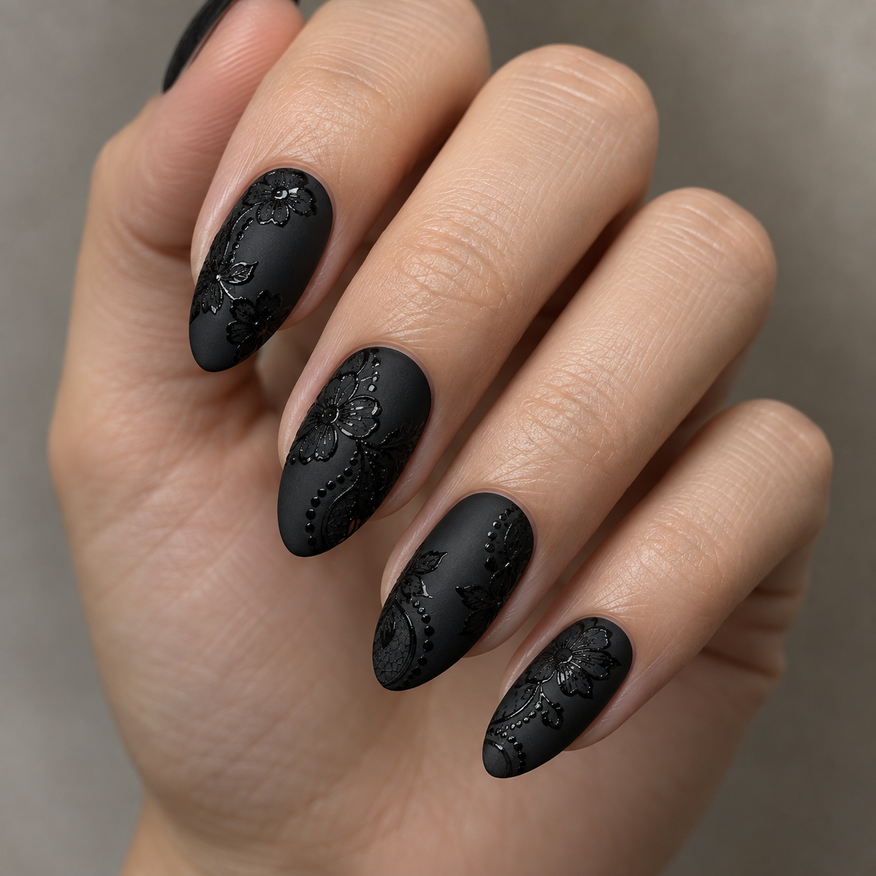 Matte black almond nails featuring glossy, raised floral and lace patterns with delicate dot accents on a neutral background.