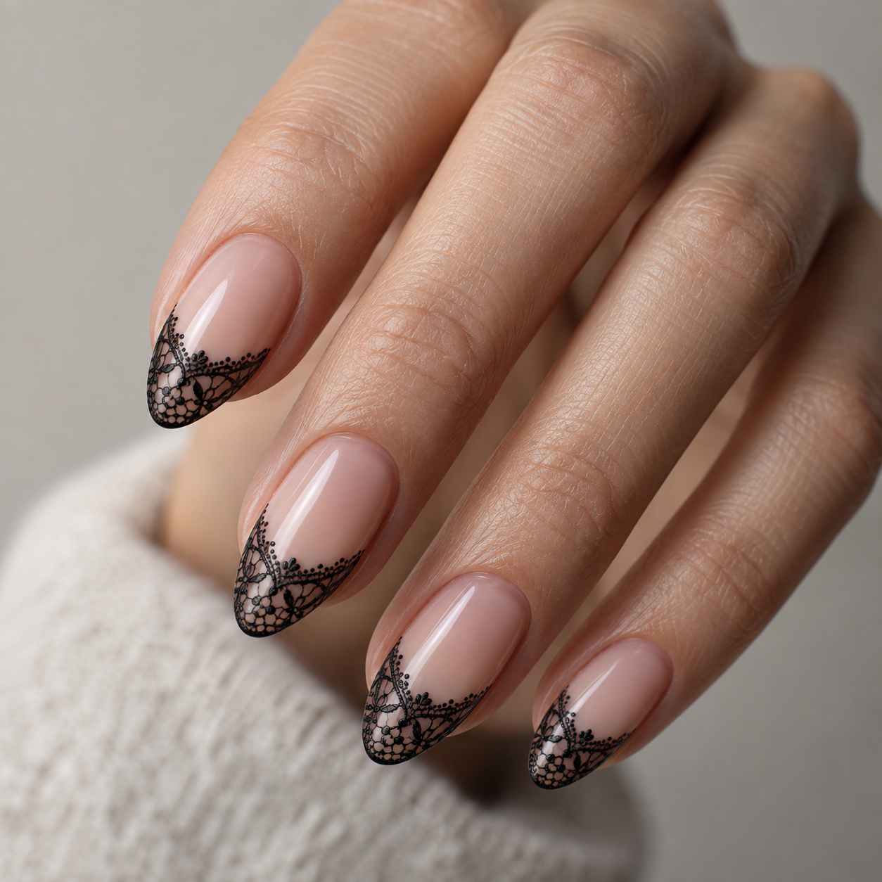 Almond nails with glossy nude base and intricate black lace pattern at the tips, shown on a hand near a white knit sleeve.
