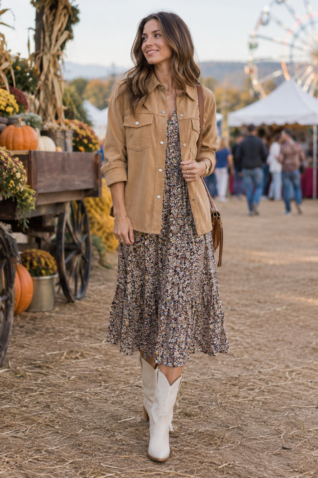 Woman in a tan suede jacket, floral midi dress, and white cowboy boots at an autumn festival. A wooden wagon with pumpkins is to her left, and a Ferris wheel stands in the background.