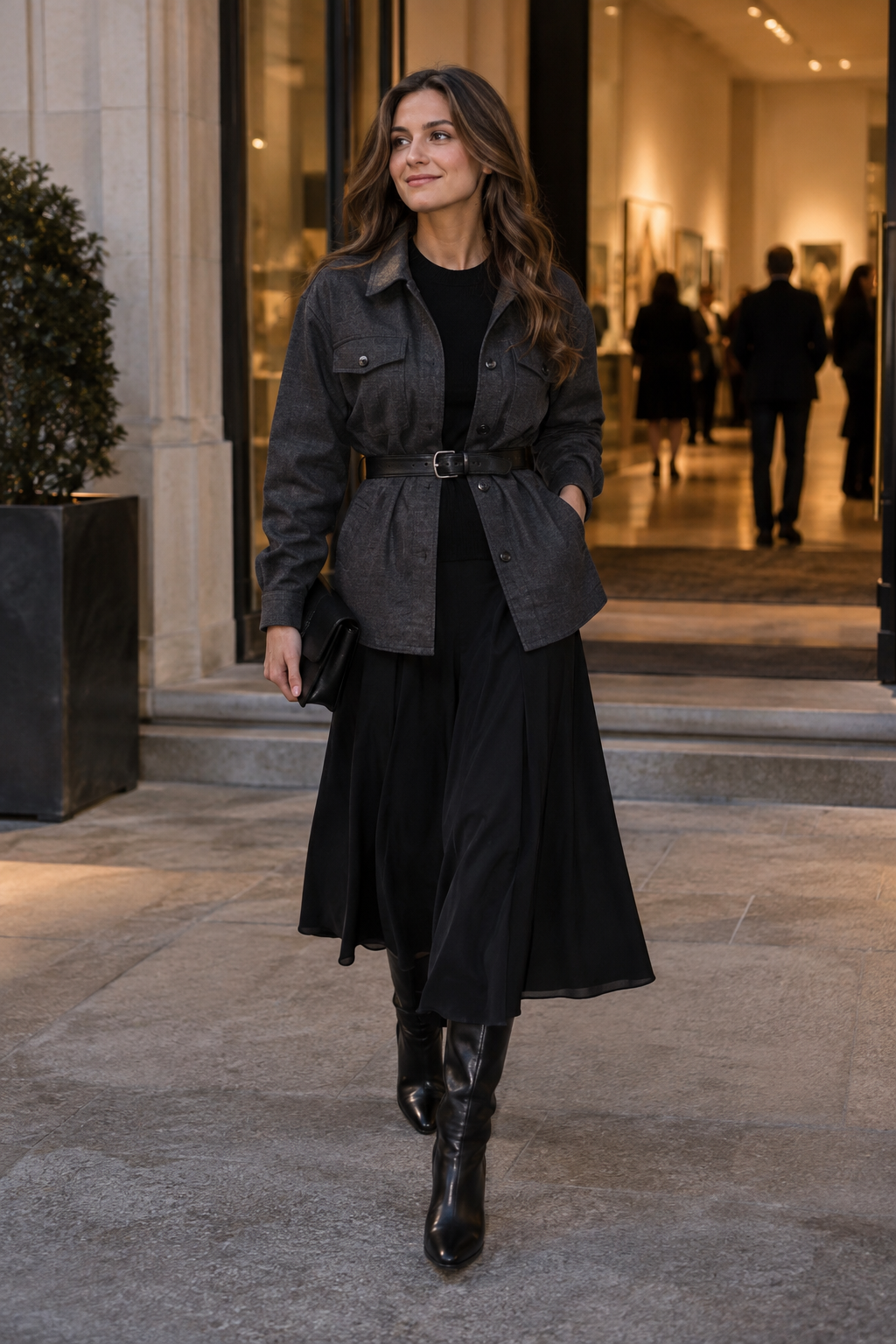 A woman walks outside wearing a belted grey jacket over a black top and midi skirt, paired with black leather boots and holding a small clutch.