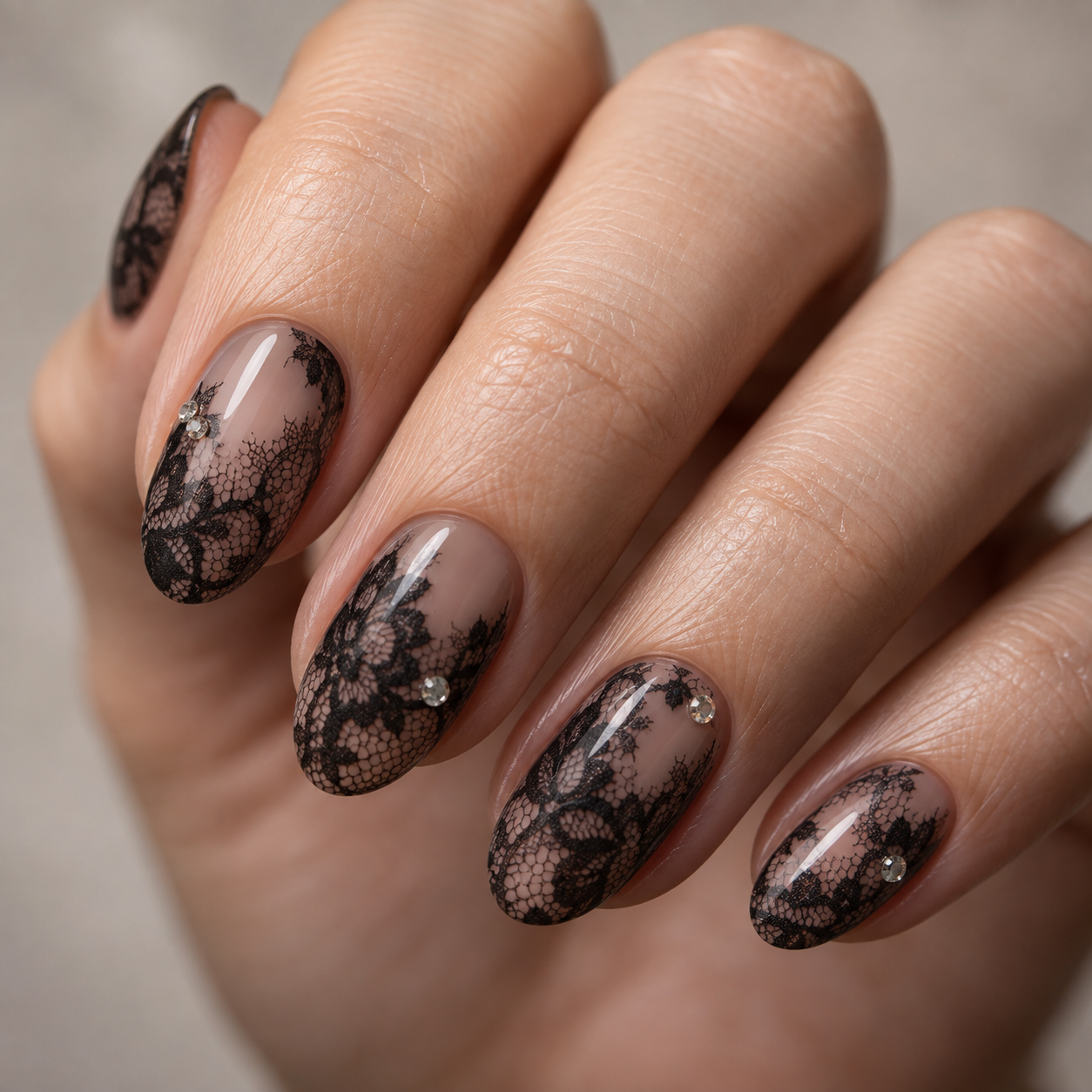 Oval nails feature a nude base with black lace overlay and small rhinestone accents near the cuticle on each visible finger.