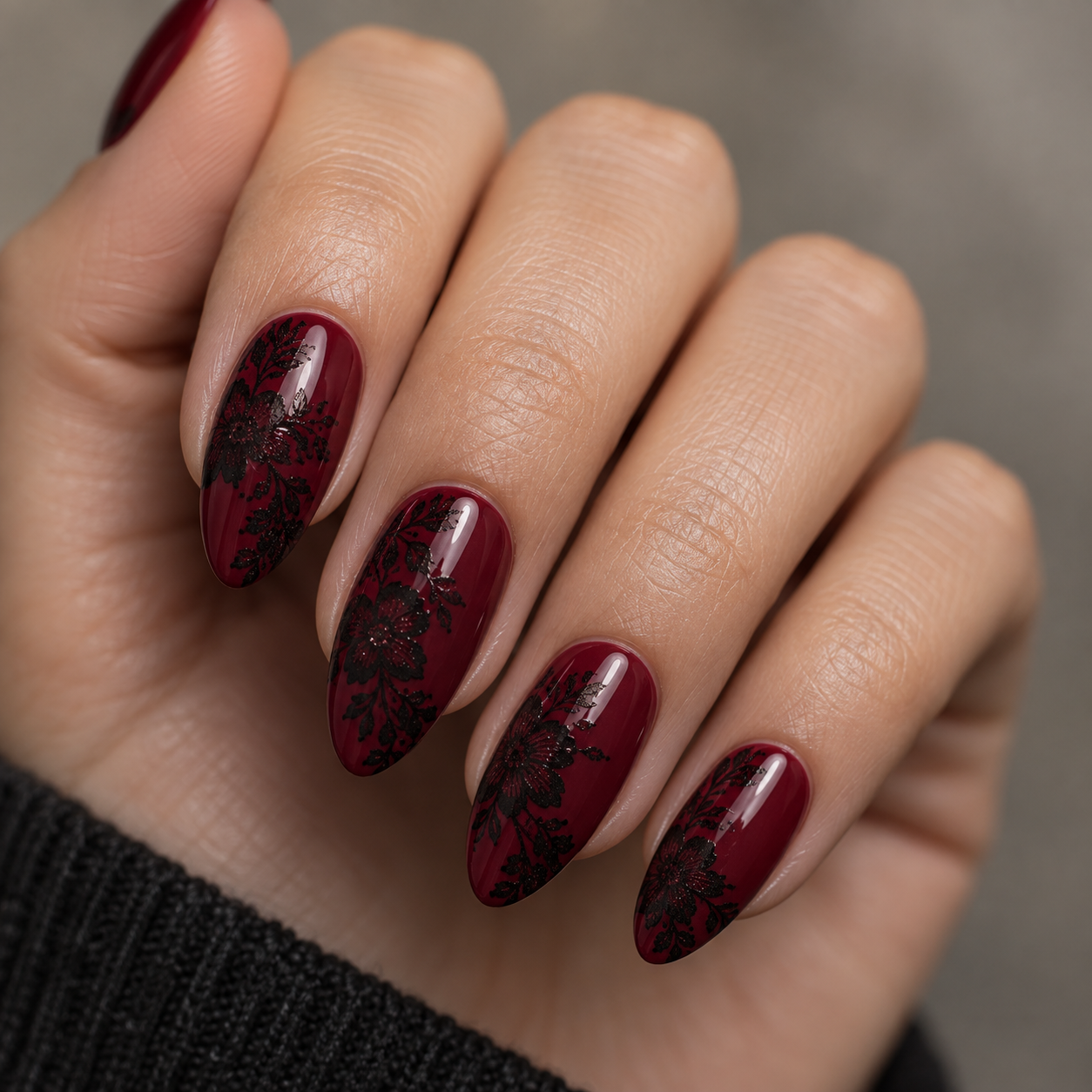 Almond-shaped nails painted deep burgundy with intricate black floral lace patterns. Glossy finish on a hand against a grey background.