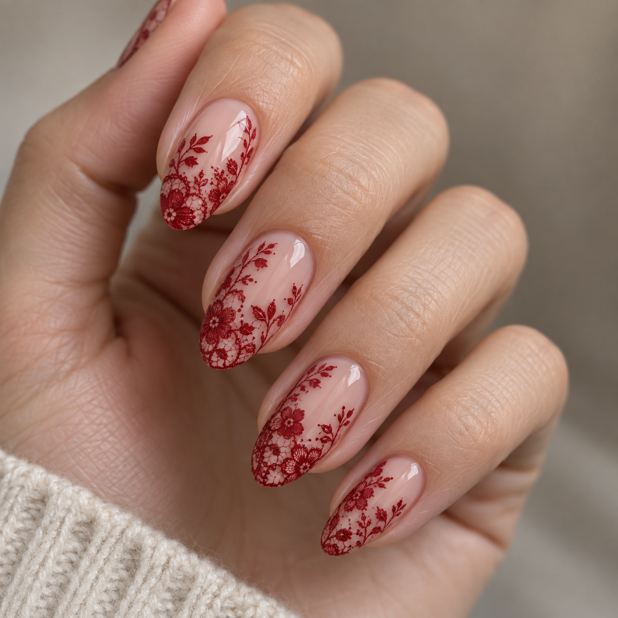 Almond nails feature a nude base with intricate red floral lace patterns. The design includes delicate flowers and vines, creating an elegant manicure look against a neutral background.