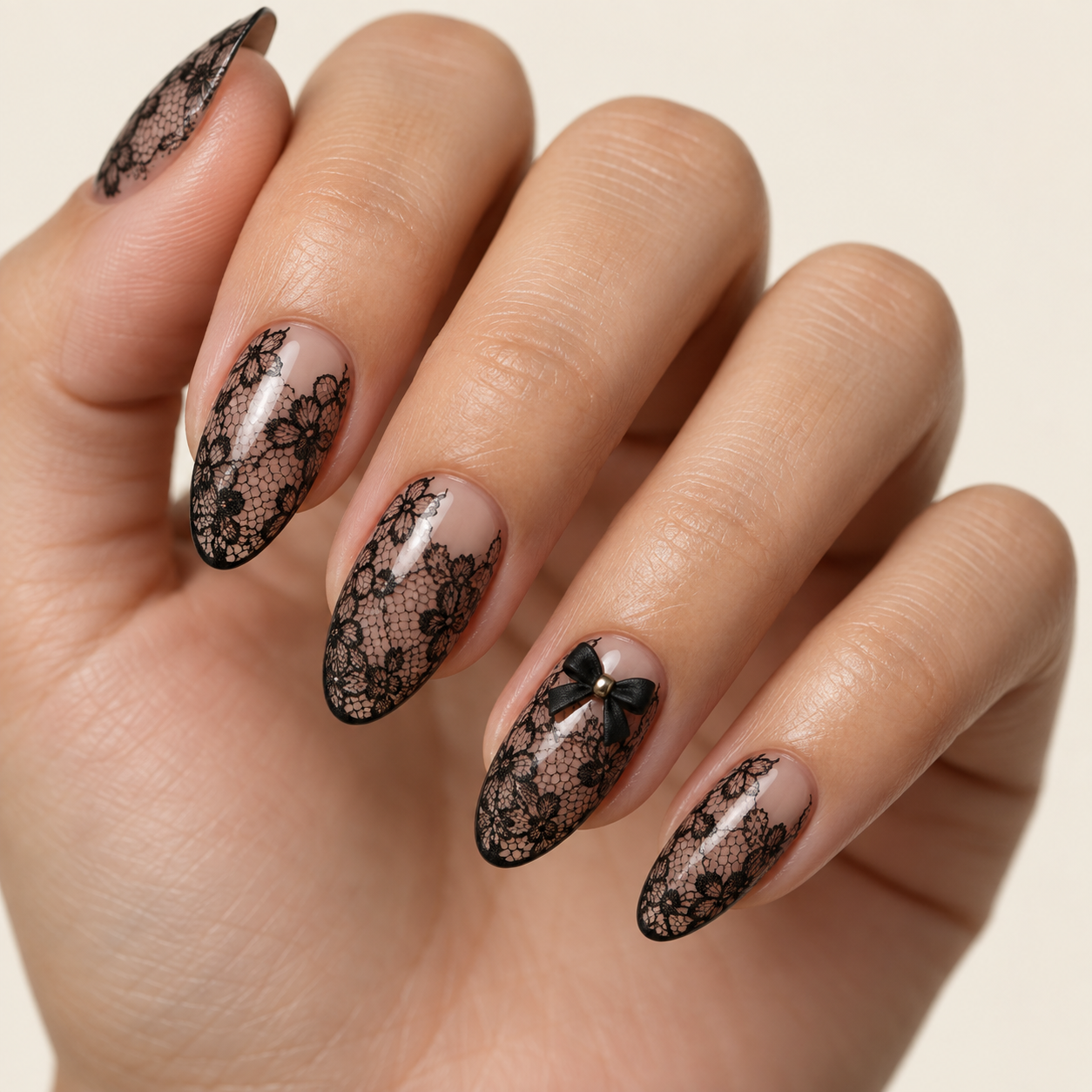 Oval nails feature black lace floral patterns on a nude base. The ring finger includes a 3D black bow with a gold center bead against a plain background.