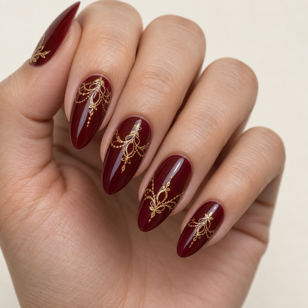 Long, almond-shaped nails painted deep burgundy with intricate gold filigree nail art on a glossy finish.
