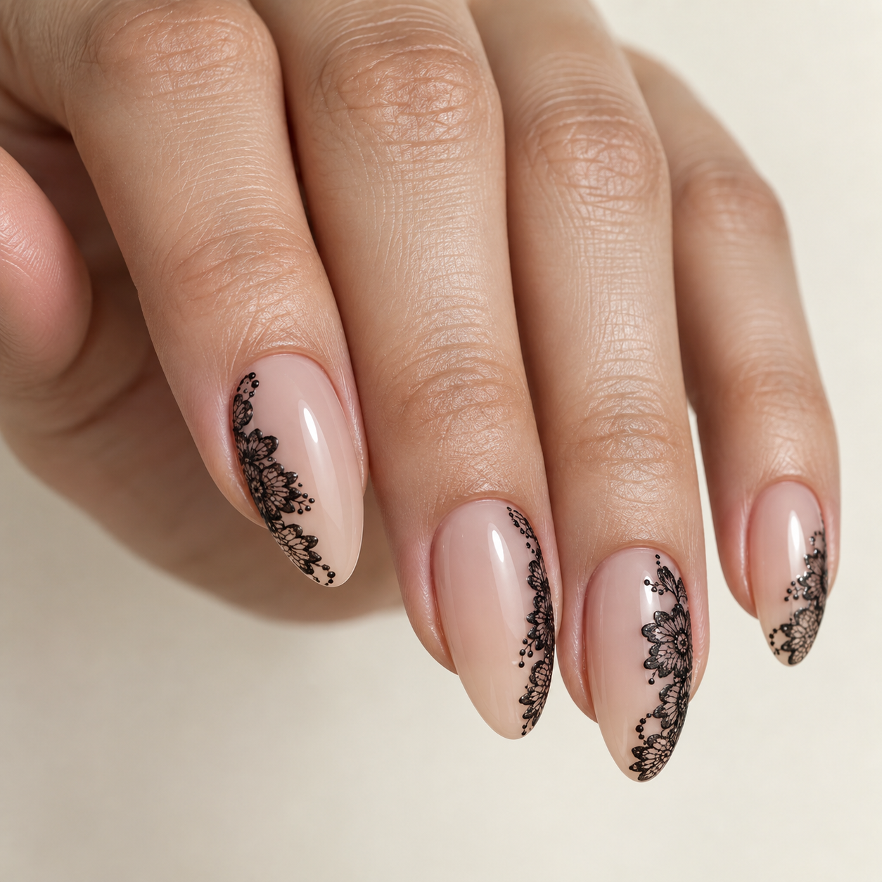 Almond-shaped nails with glossy nude polish feature intricate black lace floral patterns along the sides and tips, accented with tiny black dots.