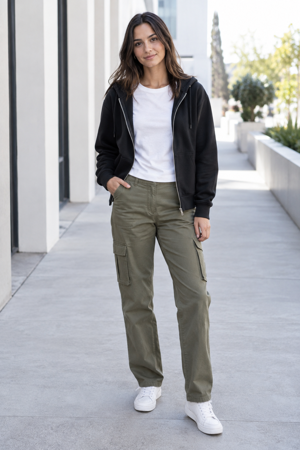 A woman stands outdoors wearing an open black zip-up hoodie over a white t-shirt, olive green cargo pants, and white sneakers, with one hand in her pocket.