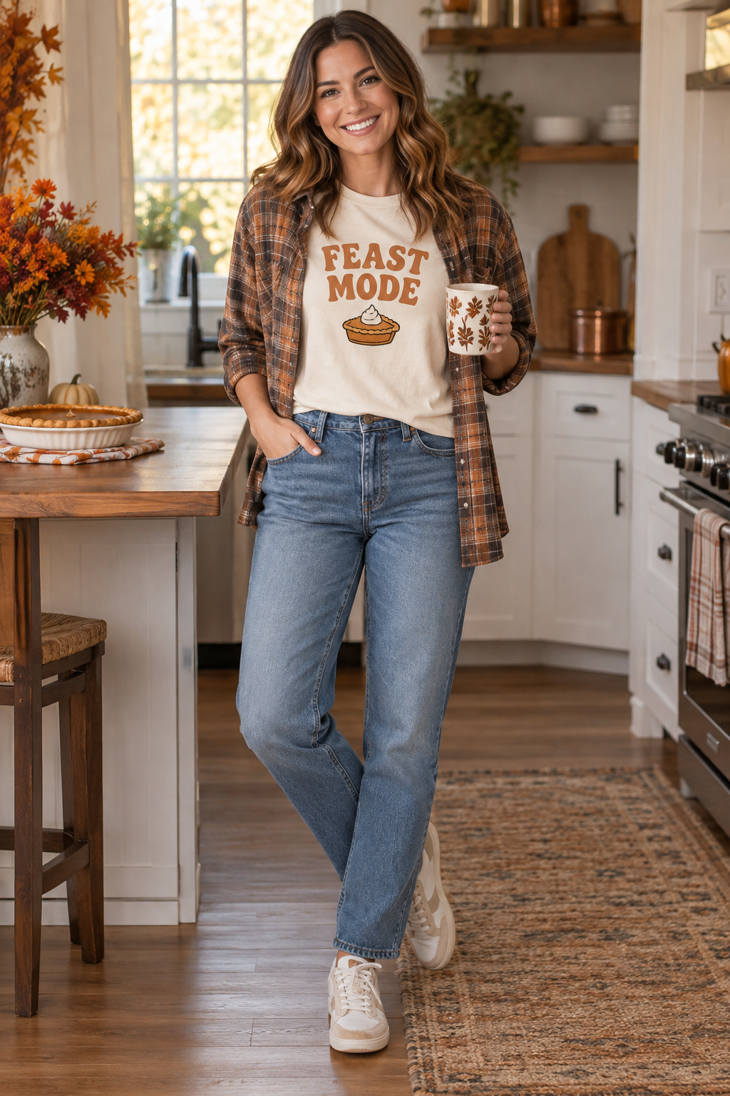 Smiling woman in a kitchen wears a "FEAST MODE" t-shirt with a pie graphic, plaid shirt, and jeans. She holds a leaf-patterned mug near a pumpkin pie and fall flowers.