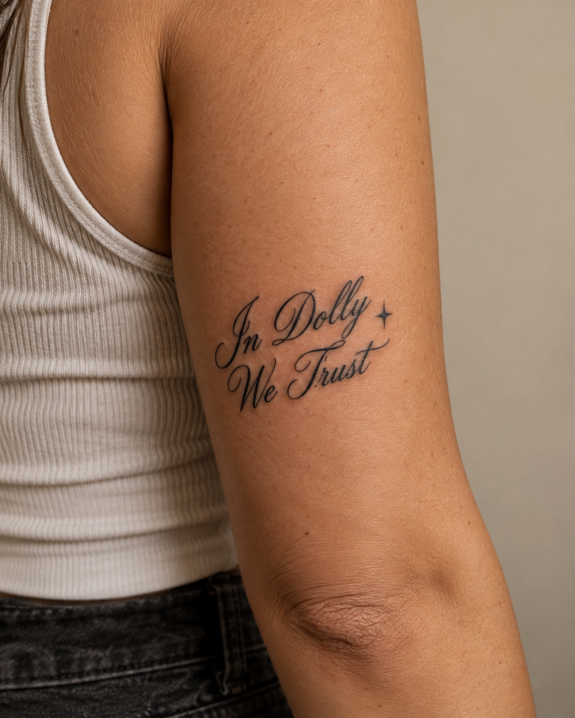 Close-up of an upper arm tattoo in black script reading "In Dolly We Trust," accompanied by a small star, on a person wearing a white ribbed tank top.