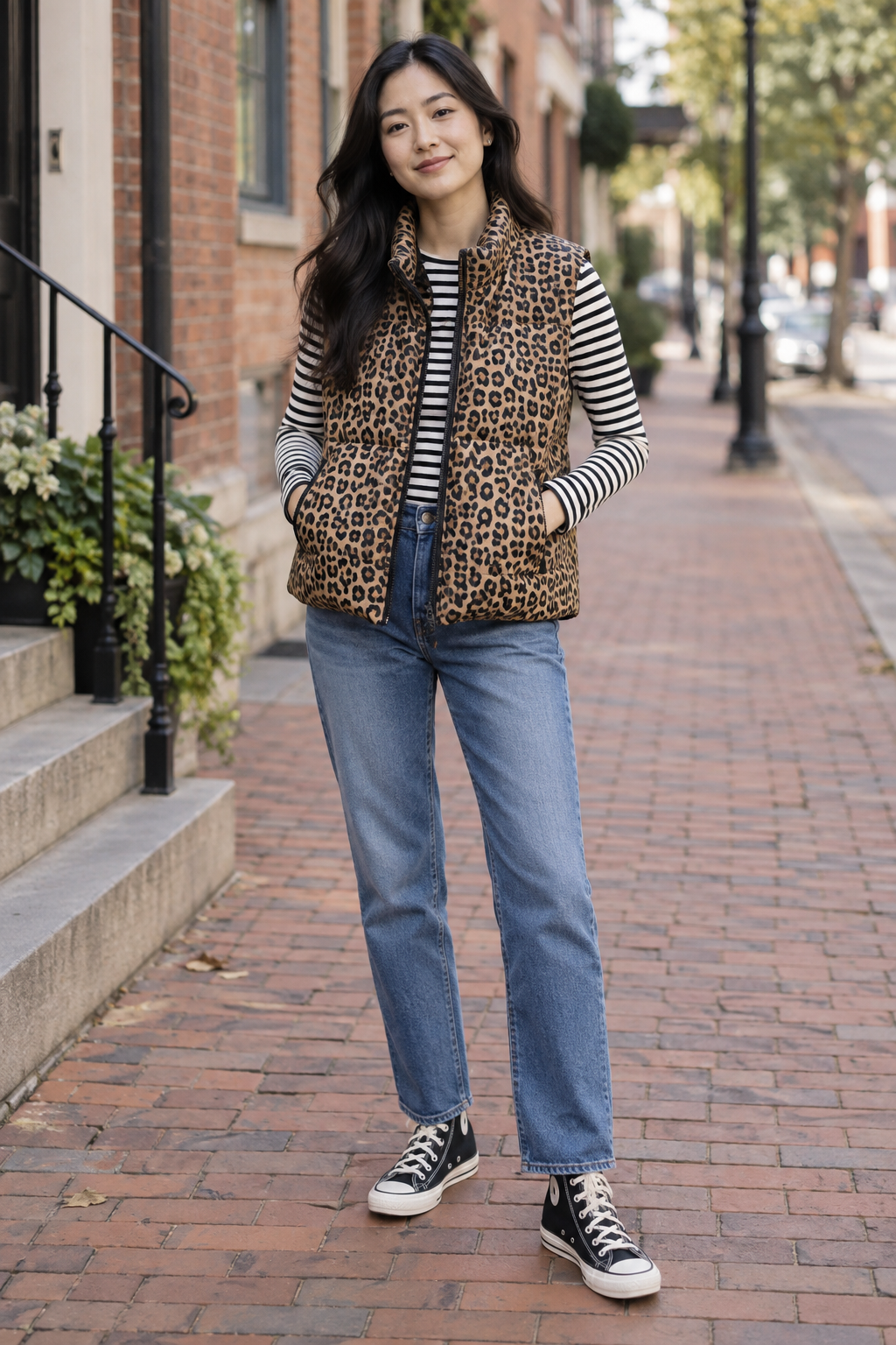 Woman with long dark hair wearing a leopard print puffer vest over a black and white striped shirt, blue jeans, and black high-top sneakers on a brick sidewalk.