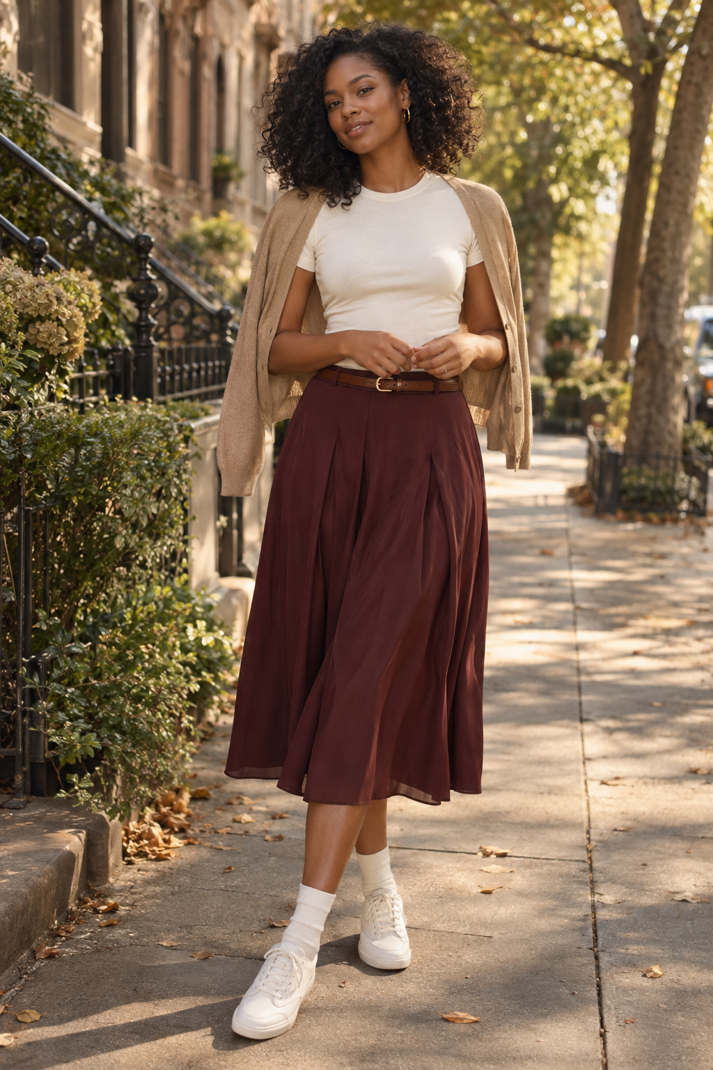 A woman with curly hair stands on a sidewalk wearing a white tee, beige cardigan draped over shoulders, maroon pleated midi skirt, brown belt, white socks, and white sneakers.