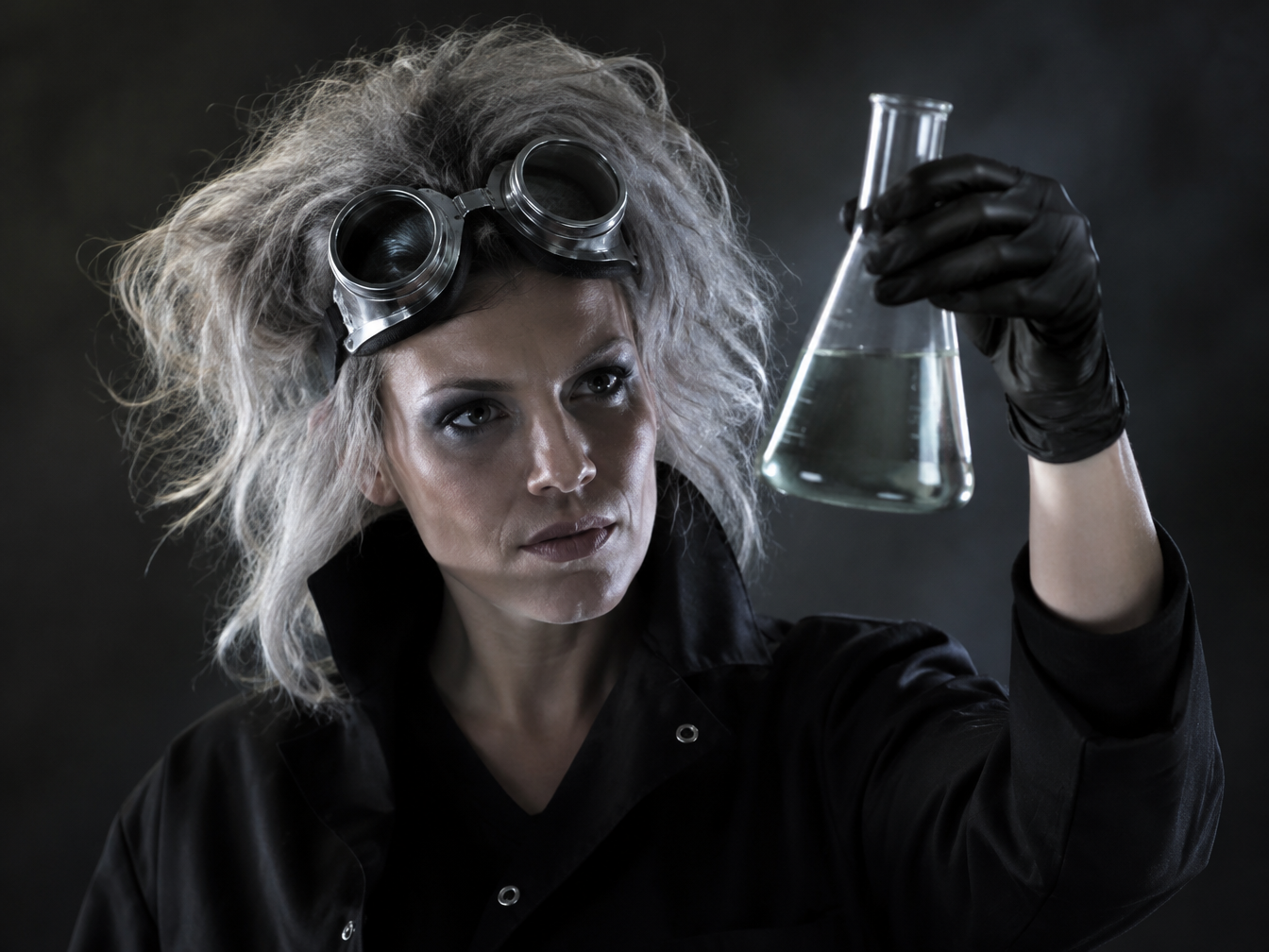 Woman in a black lab coat with popped collar, chunky goggles, nitrile gloves and grey teased hair holding a flask