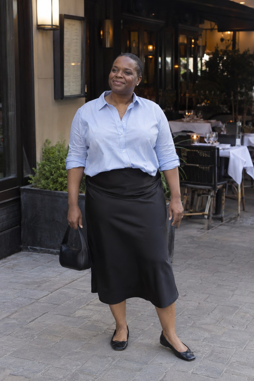 Woman in a pale blue shirt and black satin midi skirt with black ballet flats outside a restaurant.