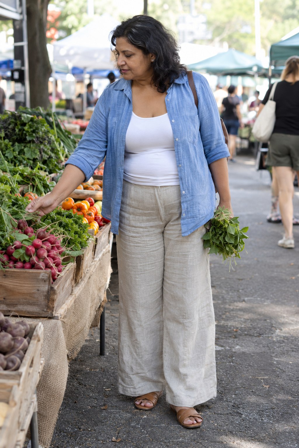 Woman in an open blue linen shirt, white tank, oatmeal linen trousers and brown sandals at a market.
