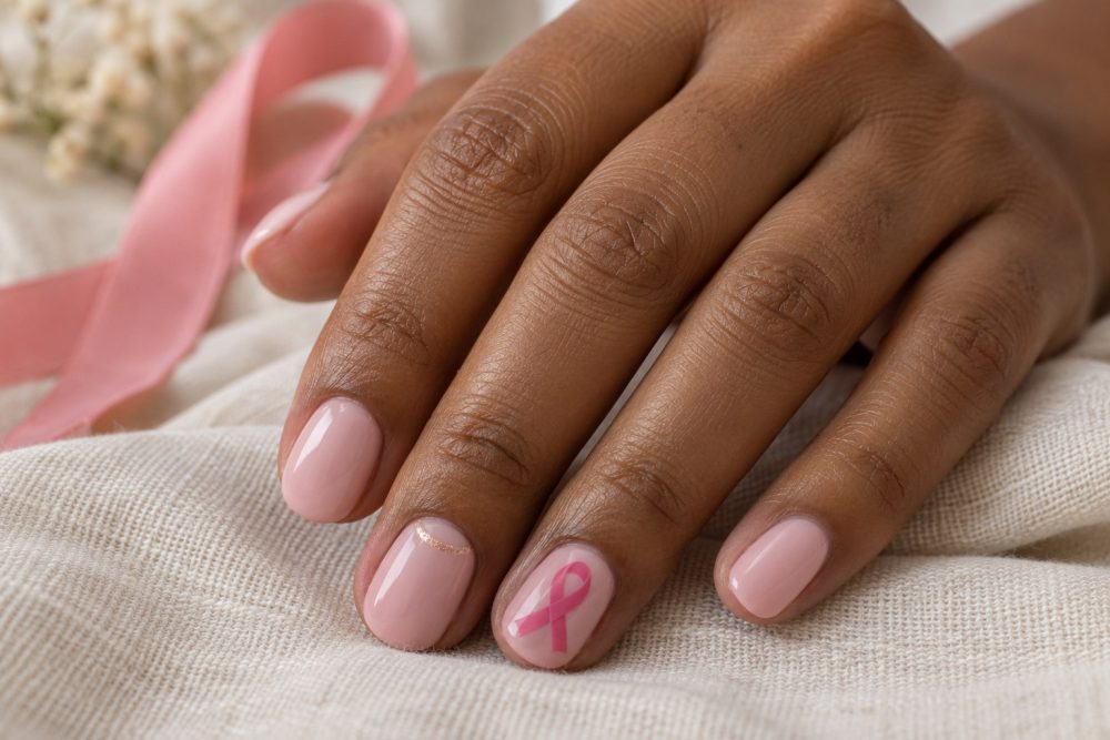 Soft pink manicure with a pink awareness ribbon and a delicate gold crescent on a linen background.