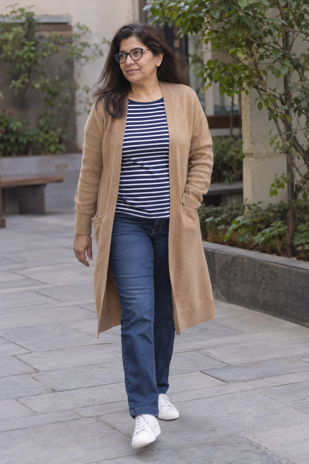 Woman wearing a long camel cardigan, striped tee, indigo straight jeans and white sneakers.