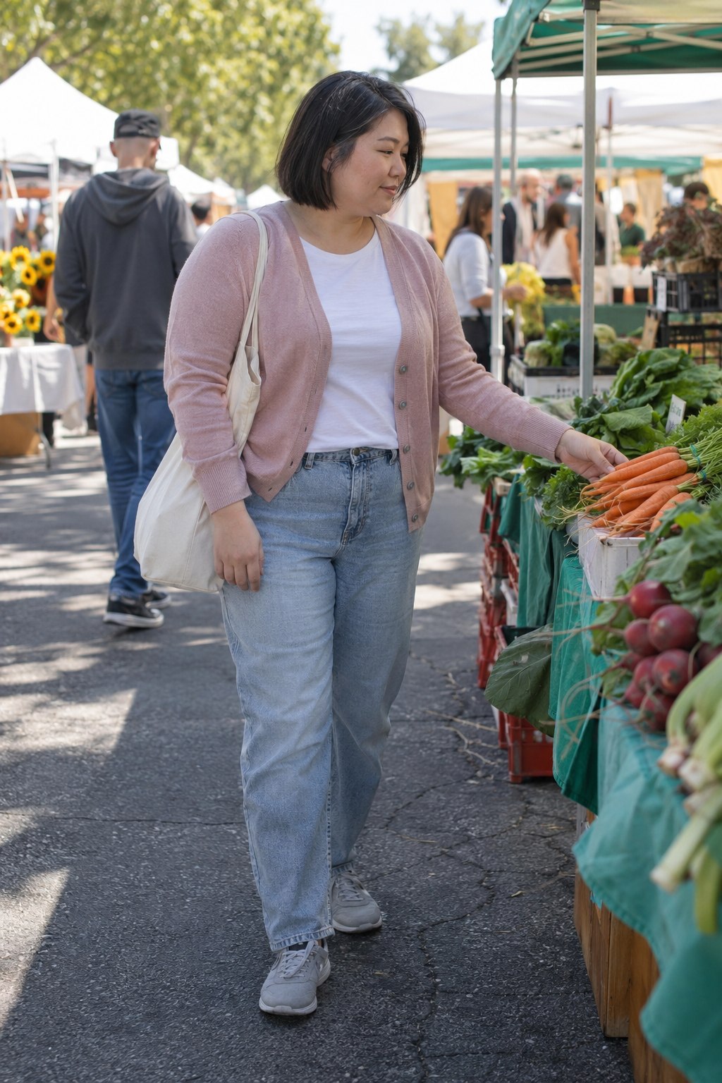 Woman wearing a pale pink cardigan, faded relaxed jeans and gray sneakers at a weekend market.