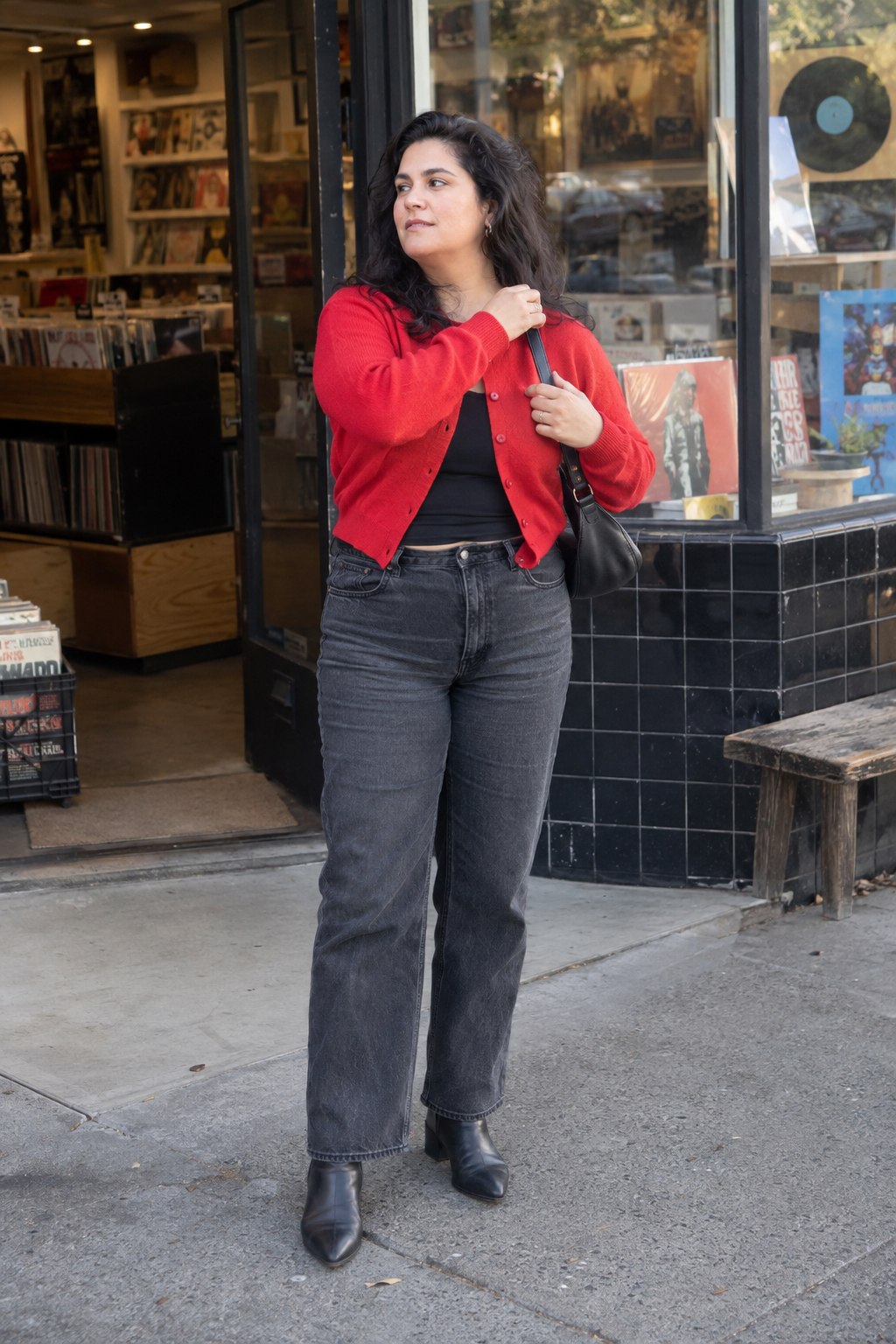 Woman in a red cardigan, washed black jeans and black ankle boots beside a record shop.
