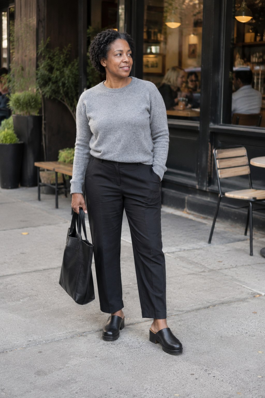 Woman wearing black chunky clogs, relaxed black trousers and a gray knit outside a cafe.
